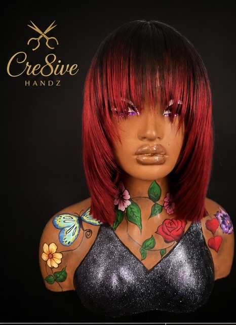 Bust of a woman with red and black hair, tattoos on shoulders and chest, wearing a black top, against a dark background.