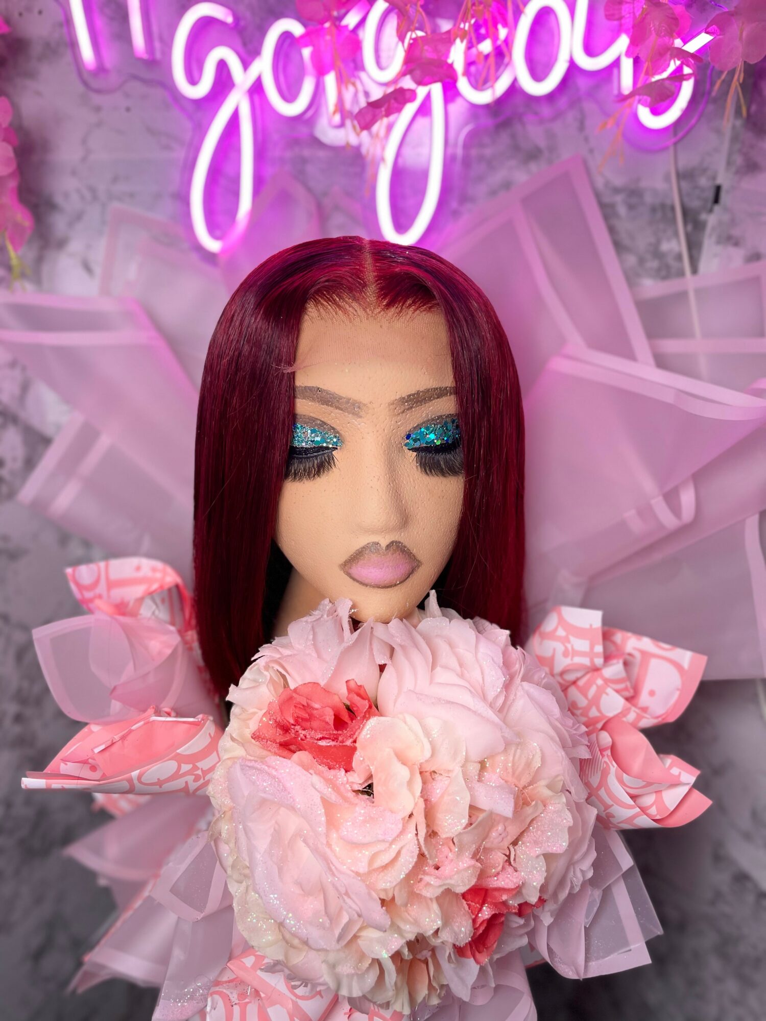Doll with long red hair, blue eyeshadow, holding pink flowers, pink background, neon sign, decorative ribbons.