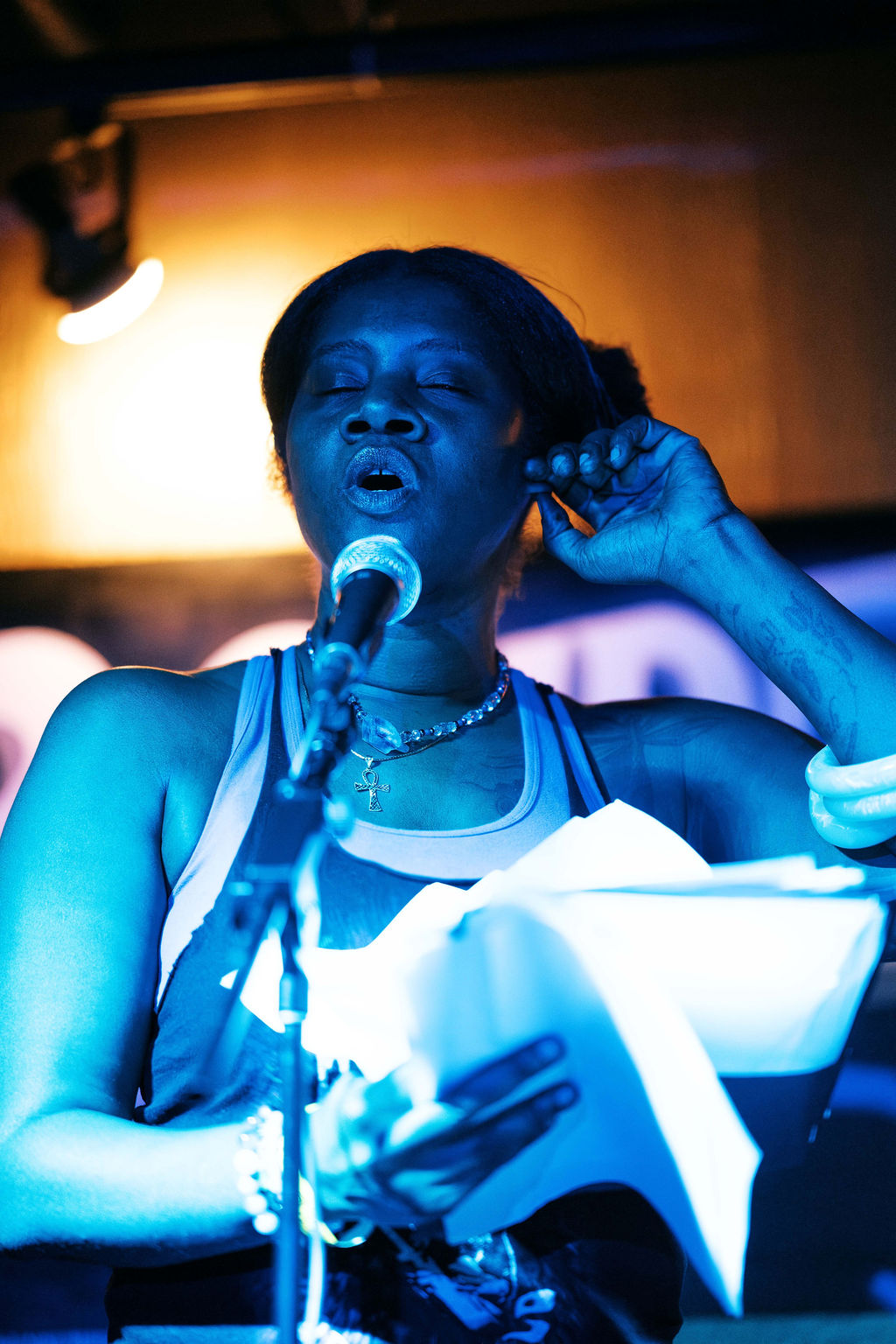 Woman singing into a microphone with eyes closed, holding a book, in a dimly lit setting with warm background light.
