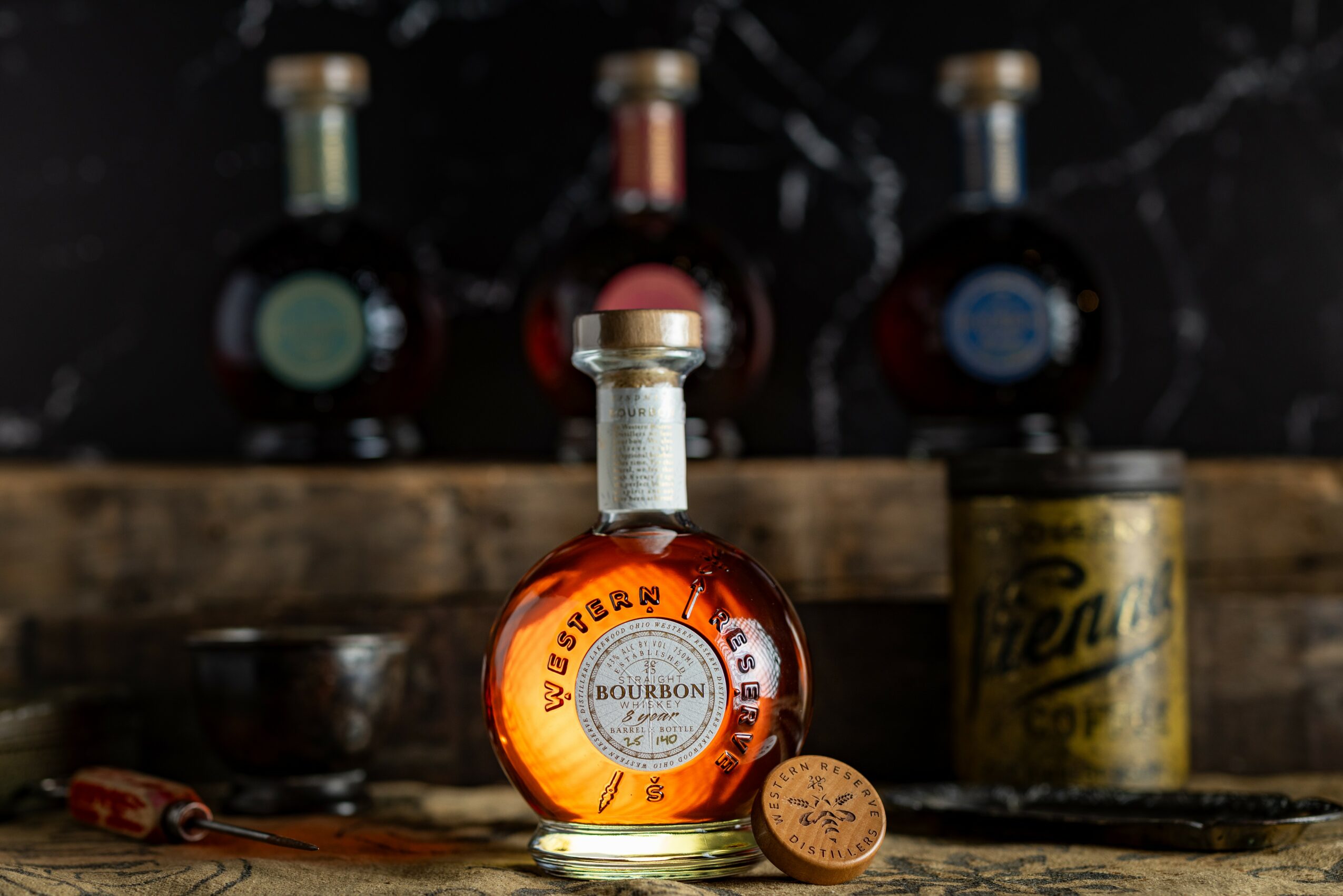 Bottle of bourbon with a round label, a cork, and a metal cap, on a wooden surface with bottles and a can in background.