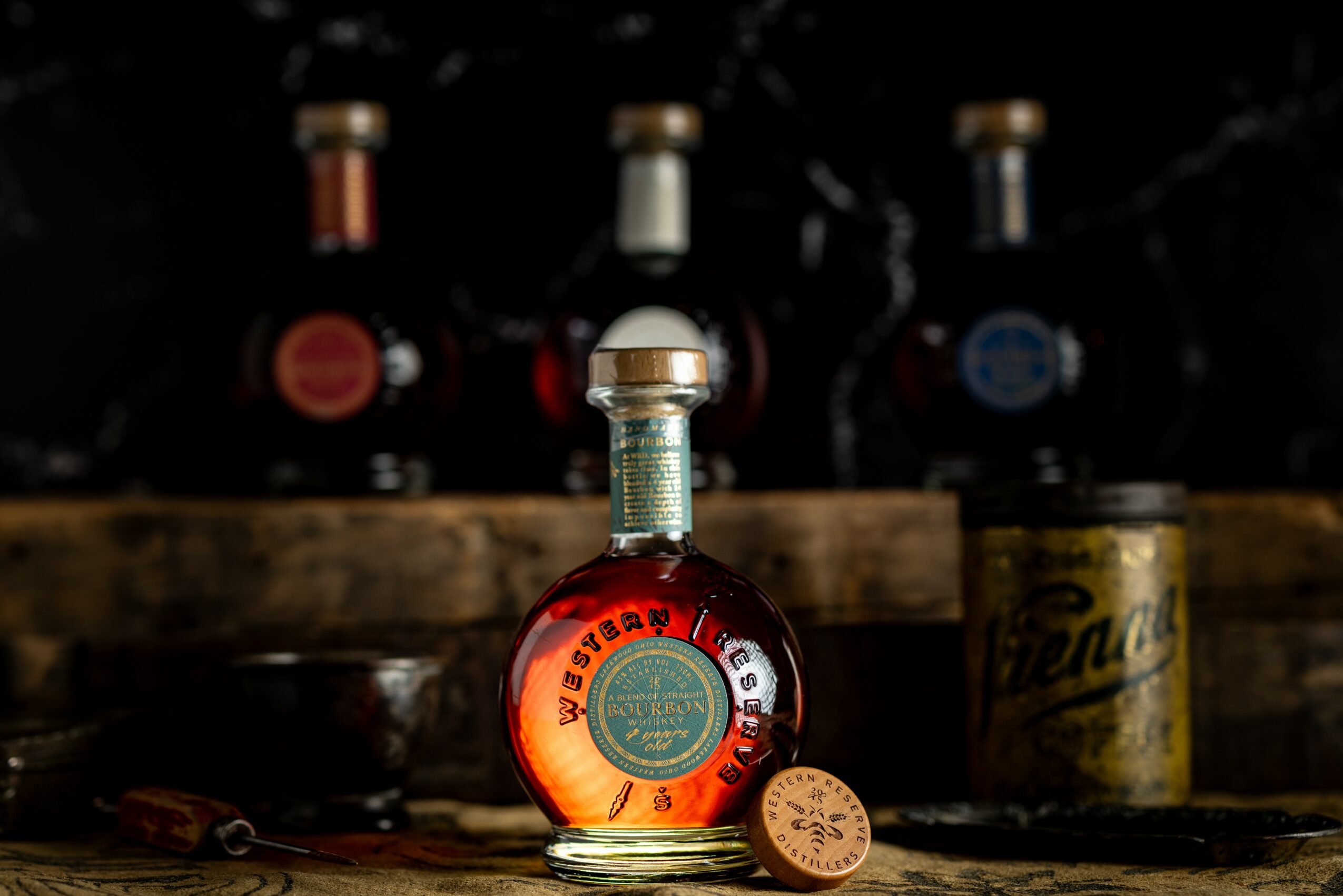 A round bottle with a gold cap and a label, placed on a wooden surface with bottles and a can in the background.