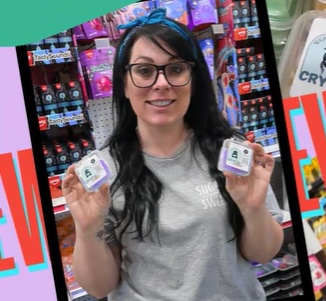 Woman with glasses holding two small containers in a store aisle with colorful products behind her.
