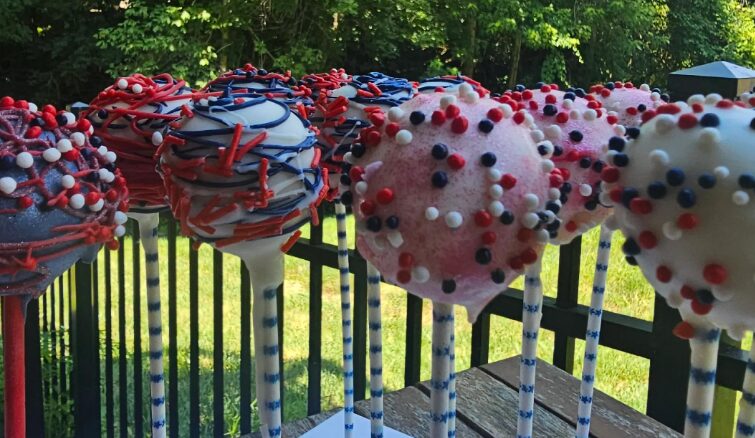 Decorated cake pops with red, white, and blue sprinkles on sticks, outdoors with a fence and trees in background.