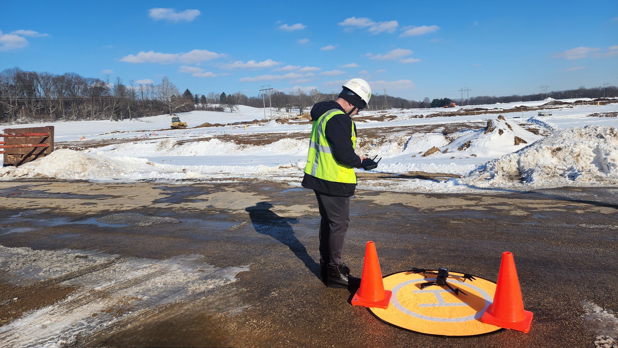 Person in safety vest and helmet standing on icy road near orange cones and a yellow device, with snow-covered landscape in background.