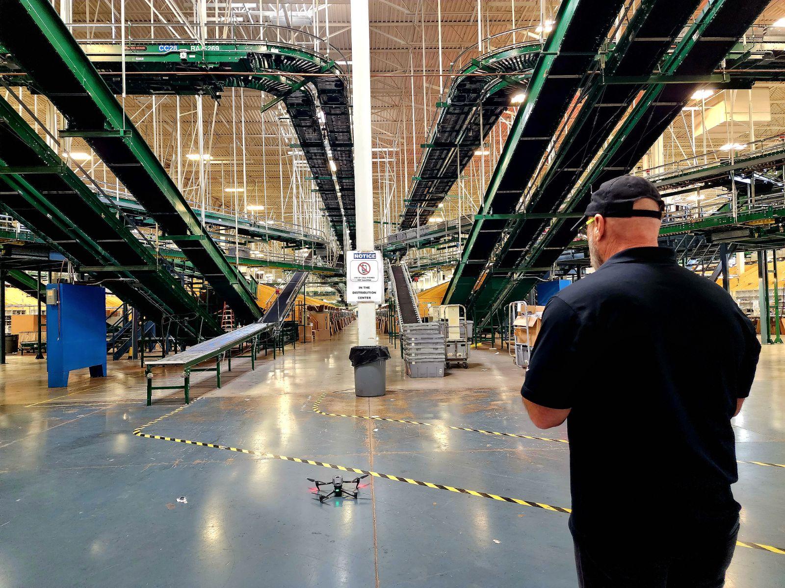 Man in black shirt and cap observing a drone on the factory floor with green pipes overhead.