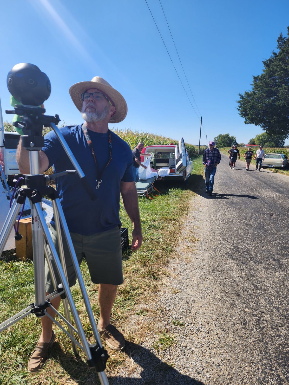 Man with a hat operating a camera on a tripod outdoors, with cars and people in the background under a clear blue sky.