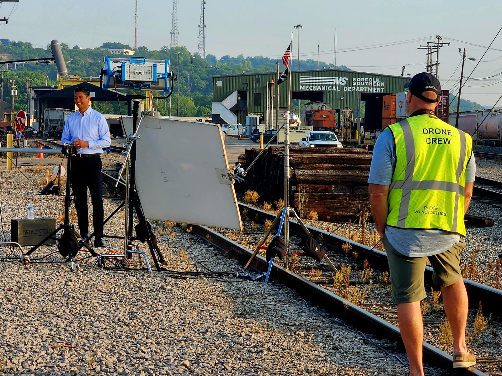 Two men working near train tracks, one with a drone crew vest, and equipment set up on gravel.