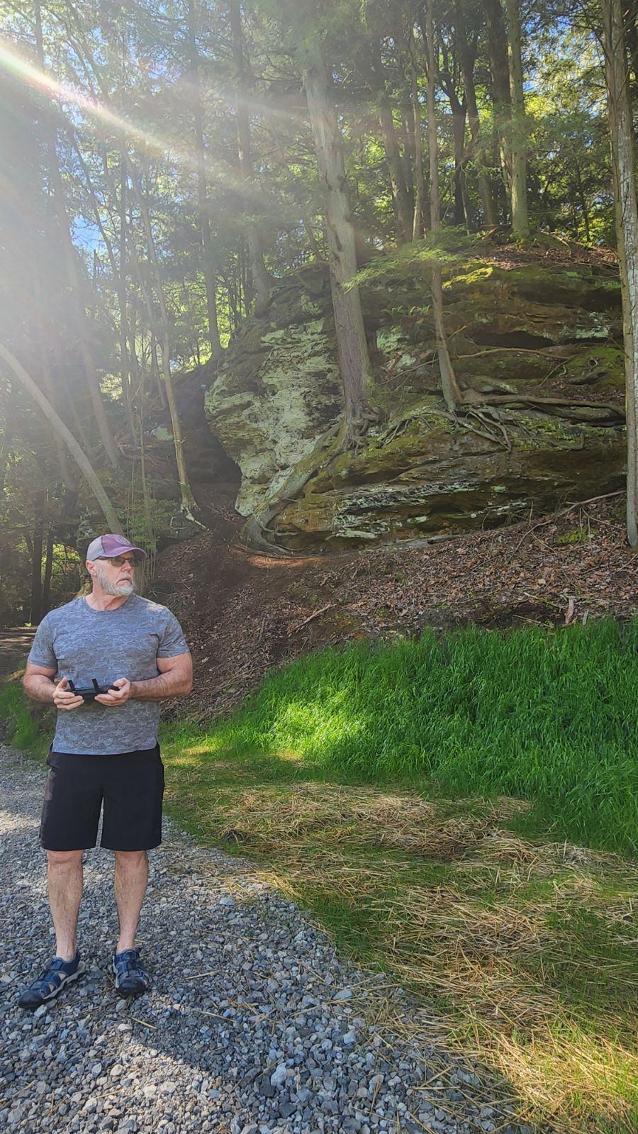 Person standing on gravel path in a forest with sunlight filtering through trees and large rocks in the background.