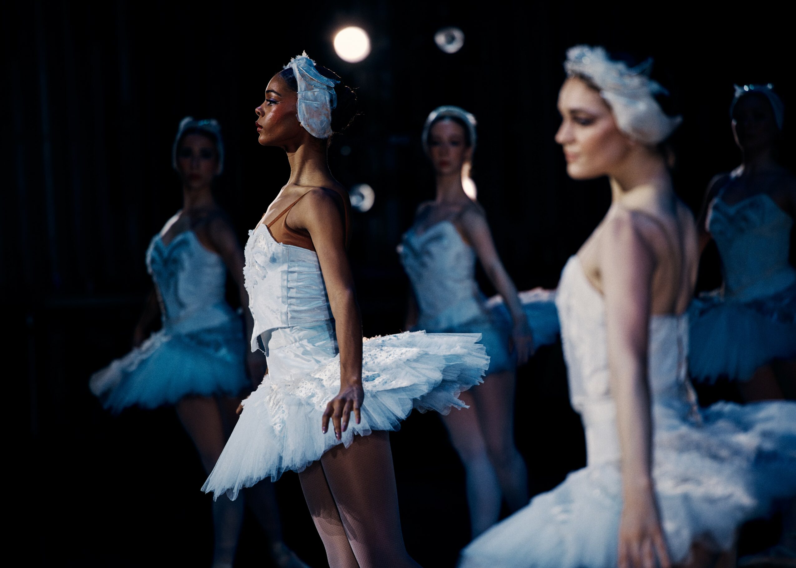 Group of ballet dancers in white tutus and headpieces on stage, illuminated by stage lights, with dark background.