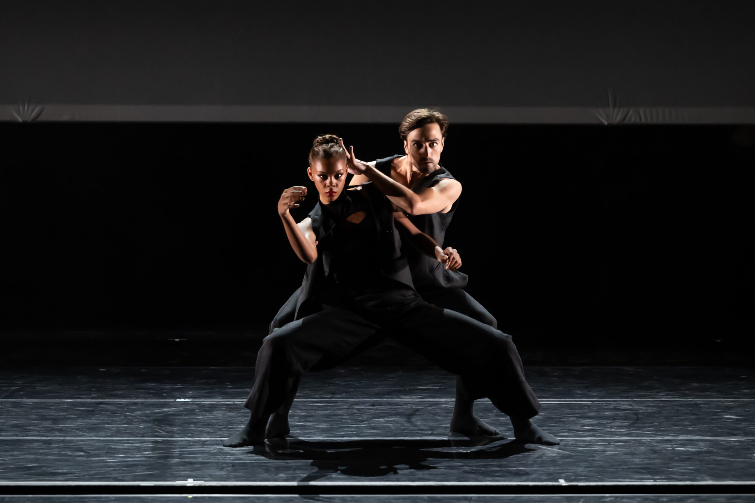 Two dancers perform on stage with a dark background, one in front and the other behind, in a contemporary dance pose.