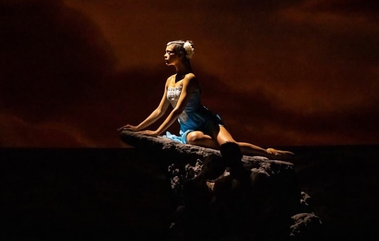 Person sitting on a rock at sunset, facing left, with a dark sky background.