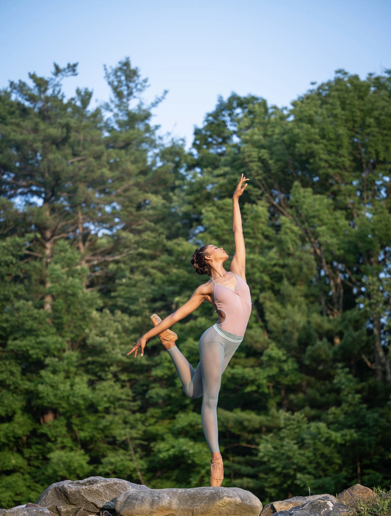 Ballet dancer performing outdoors on rocks with trees in the background, one leg raised and arms extended gracefully.