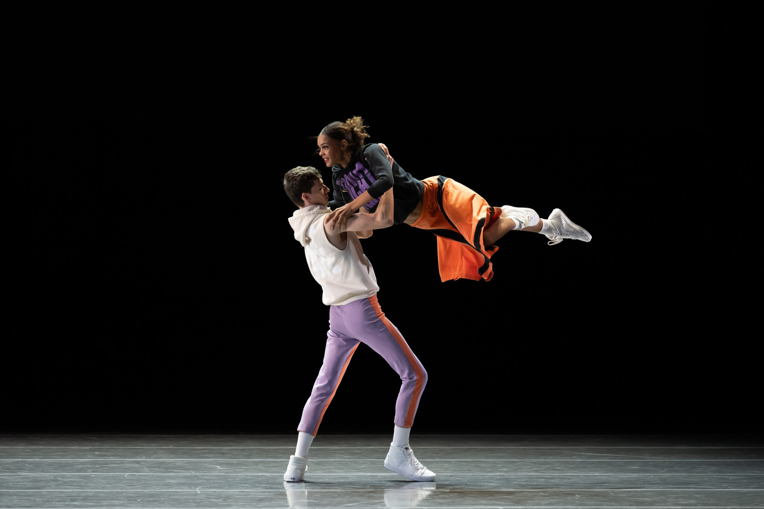 Two performers, one lifting the other, on a dark stage with a black background, engaged in a dance or acrobatic act.