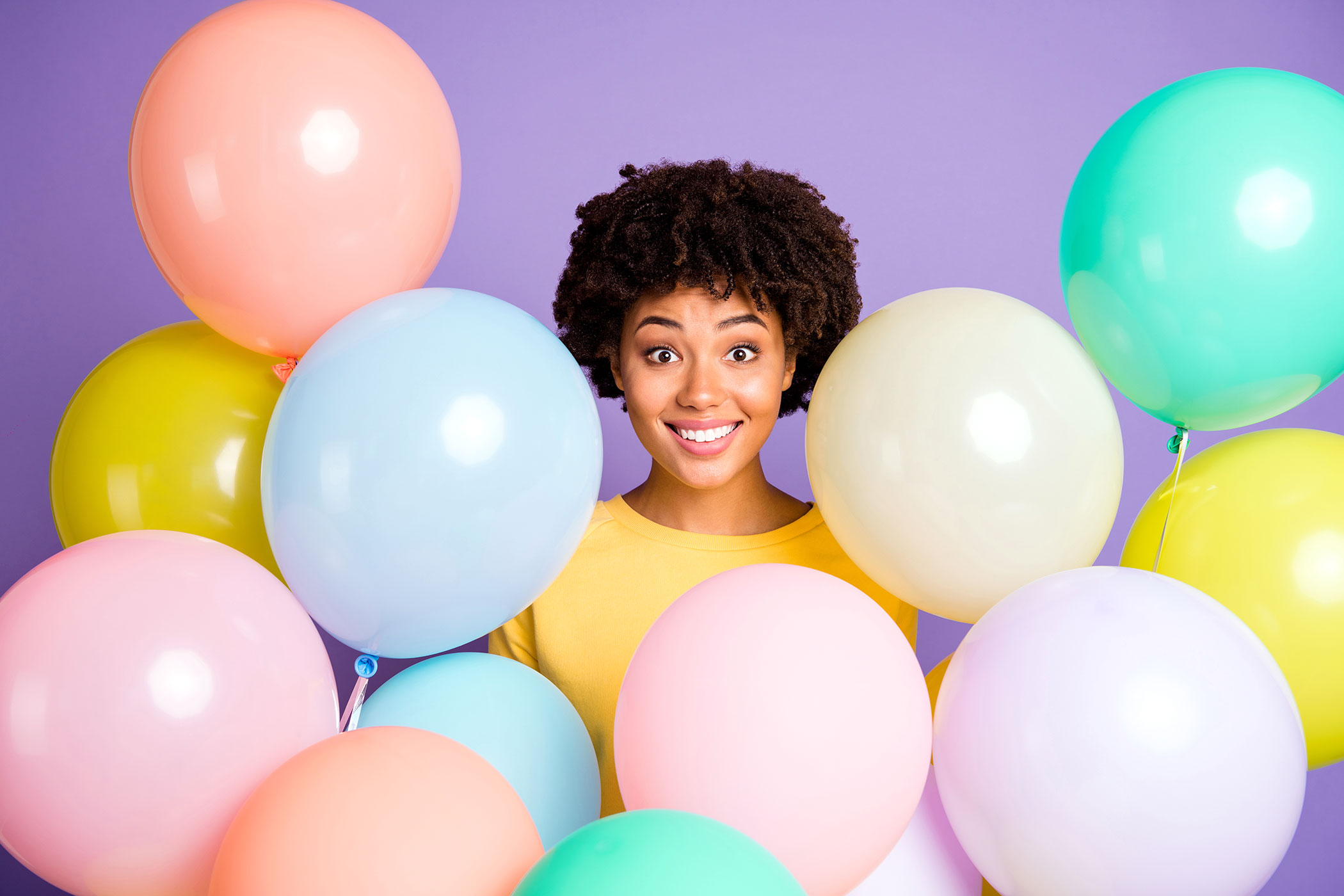Smiling woman surrounded by colorful balloons against a purple background.