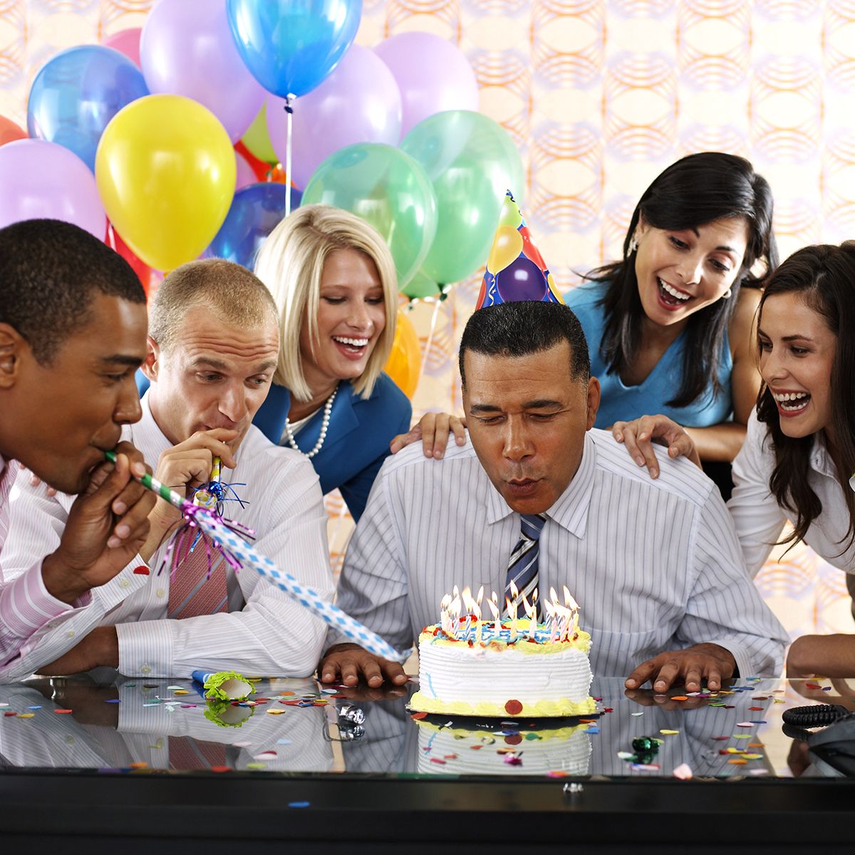 Group of people celebrating birthday with a cake, balloons, and smiling faces.