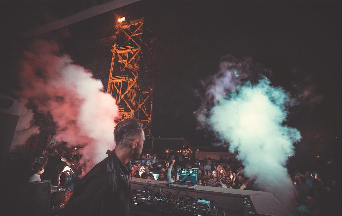 DJ performing at a concert with colorful smoke effects and a crowd in the background.
