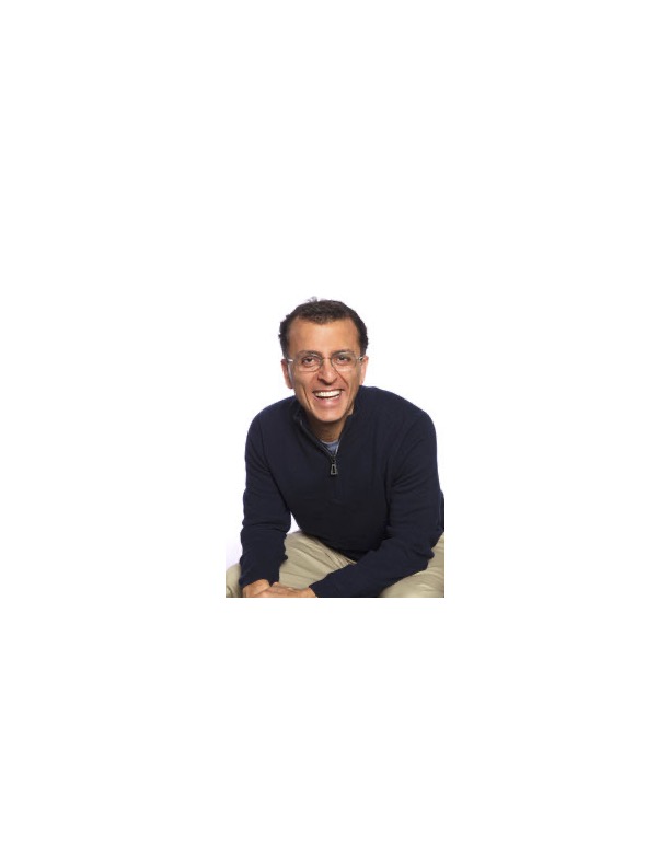Smiling man with glasses wearing a dark sweater and light-colored pants, sitting and leaning forward.