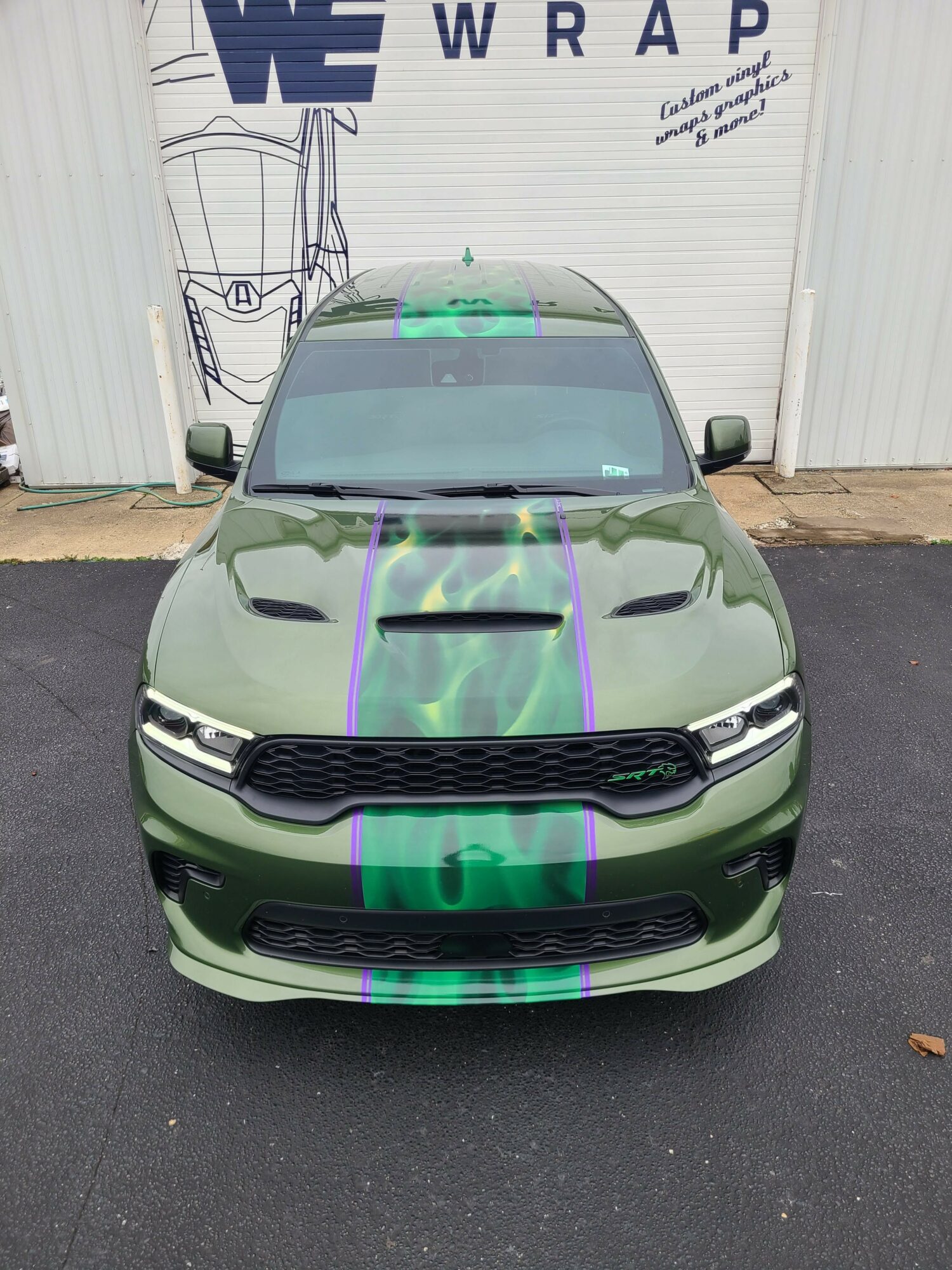 Green car with black grille and headlights parked on asphalt in front of a white building with blue text.