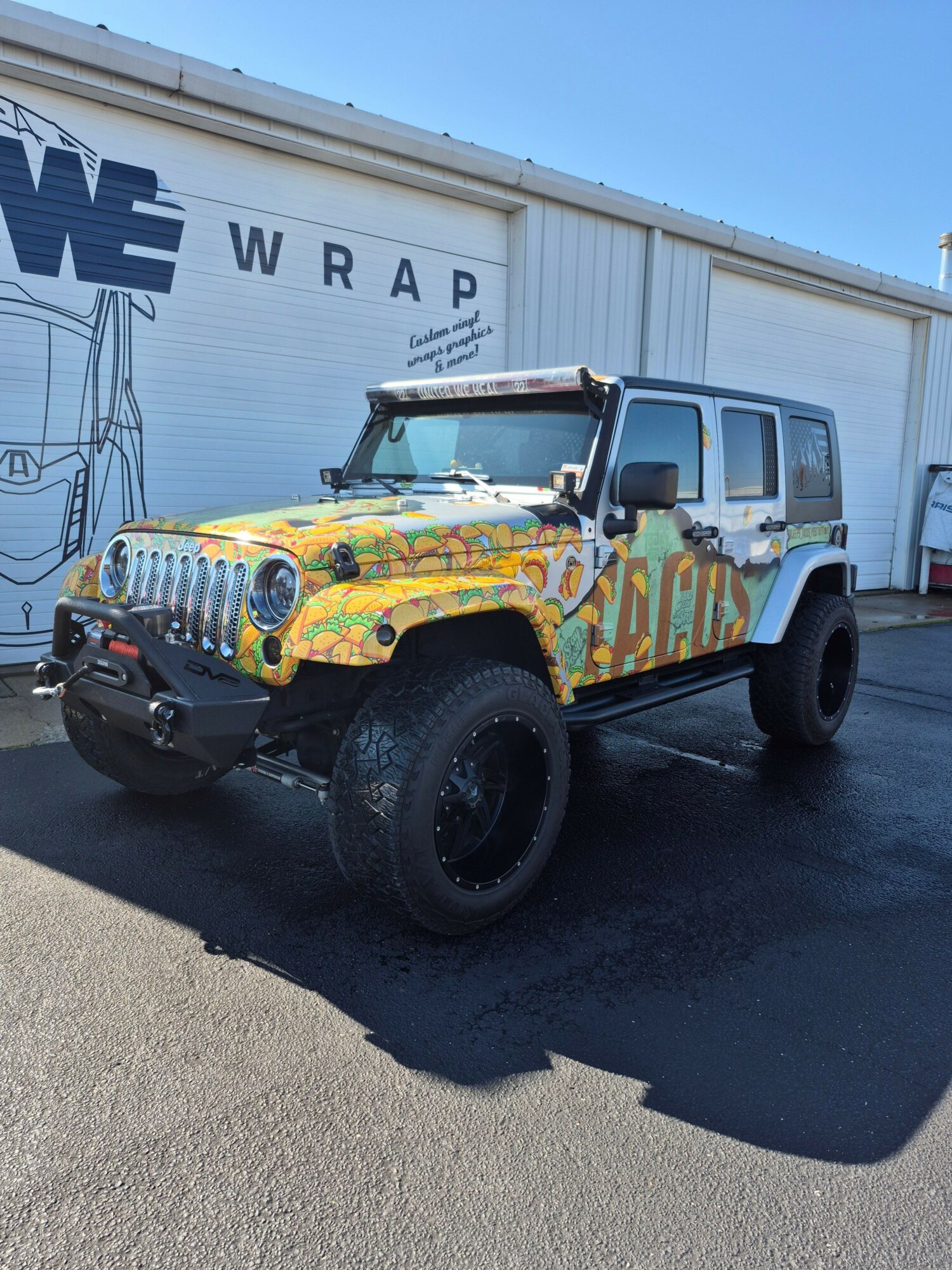 Colorful, patterned vehicle with large tires parked outside a building with a sign reading 'WRAP'.