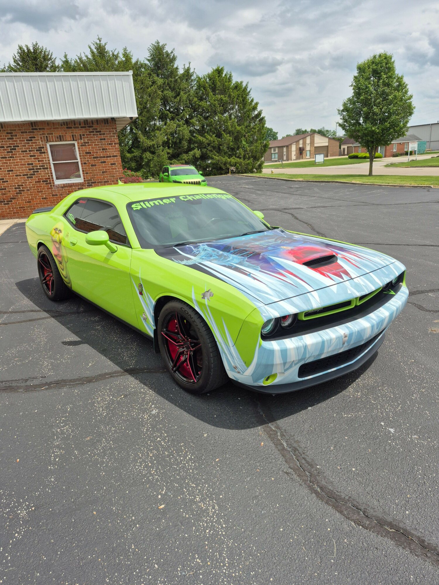 Green sports car with a colorful design on the hood parked on a street, trees and buildings in the background.