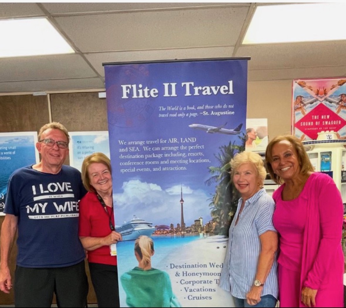 Four people standing indoors in front of a travel banner, smiling, with ceiling tiles visible above.
