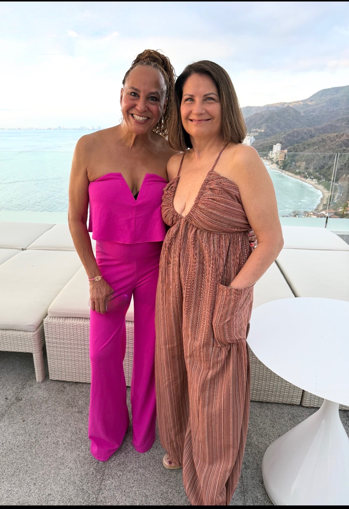 Two women standing outdoors with a coastal landscape in the background, smiling, one in a pink strapless outfit, the other in a striped jumpsuit.