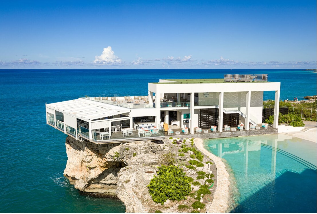 Modern building on cliff overlooking ocean with pool and clear sky, surrounded by greenery.