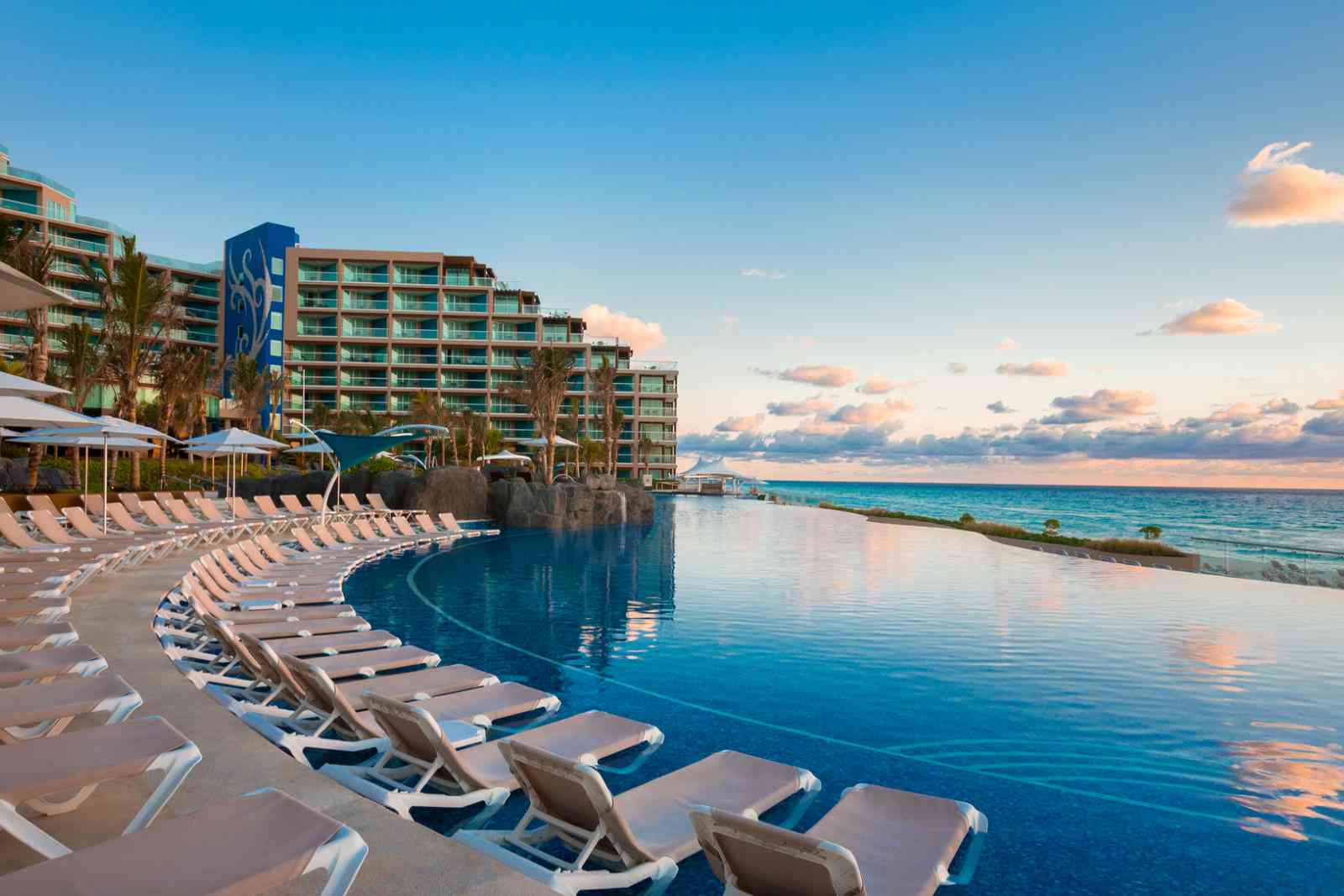 Resort with pool and beach, lounge chairs along pool, ocean view, blue sky with clouds, sunset reflection, buildings in background