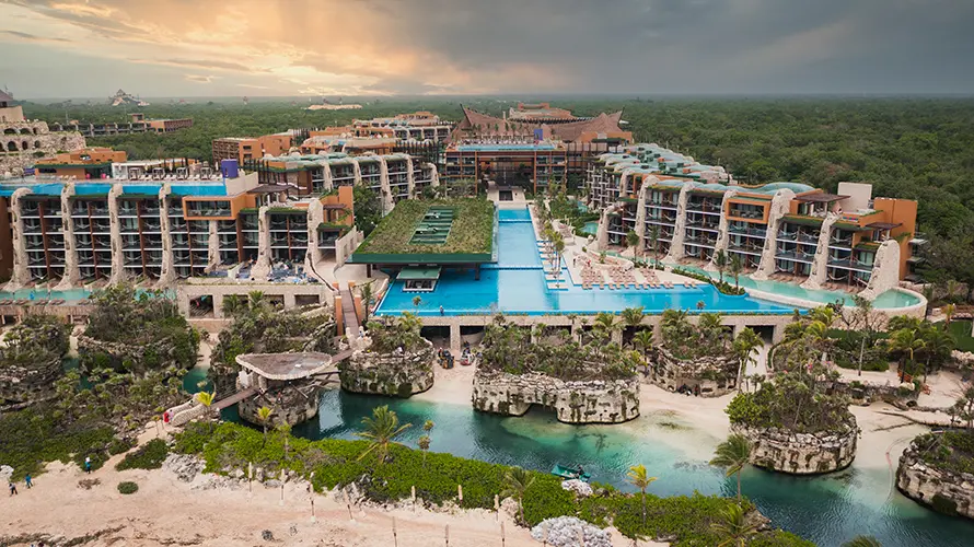 A large resort with multiple buildings, pools, and rocky landscape, surrounded by greenery and a cloudy sky.