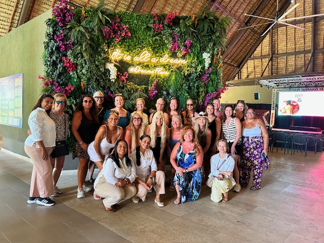 Group of women posing indoors in front of a floral wall with neon sign, some standing and some kneeling, smiling.