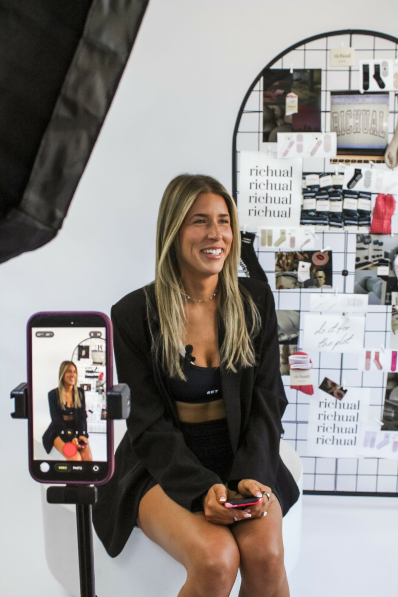 Woman sitting and smiling, holding a phone, in front of a display board with images and notes, in a bright room.