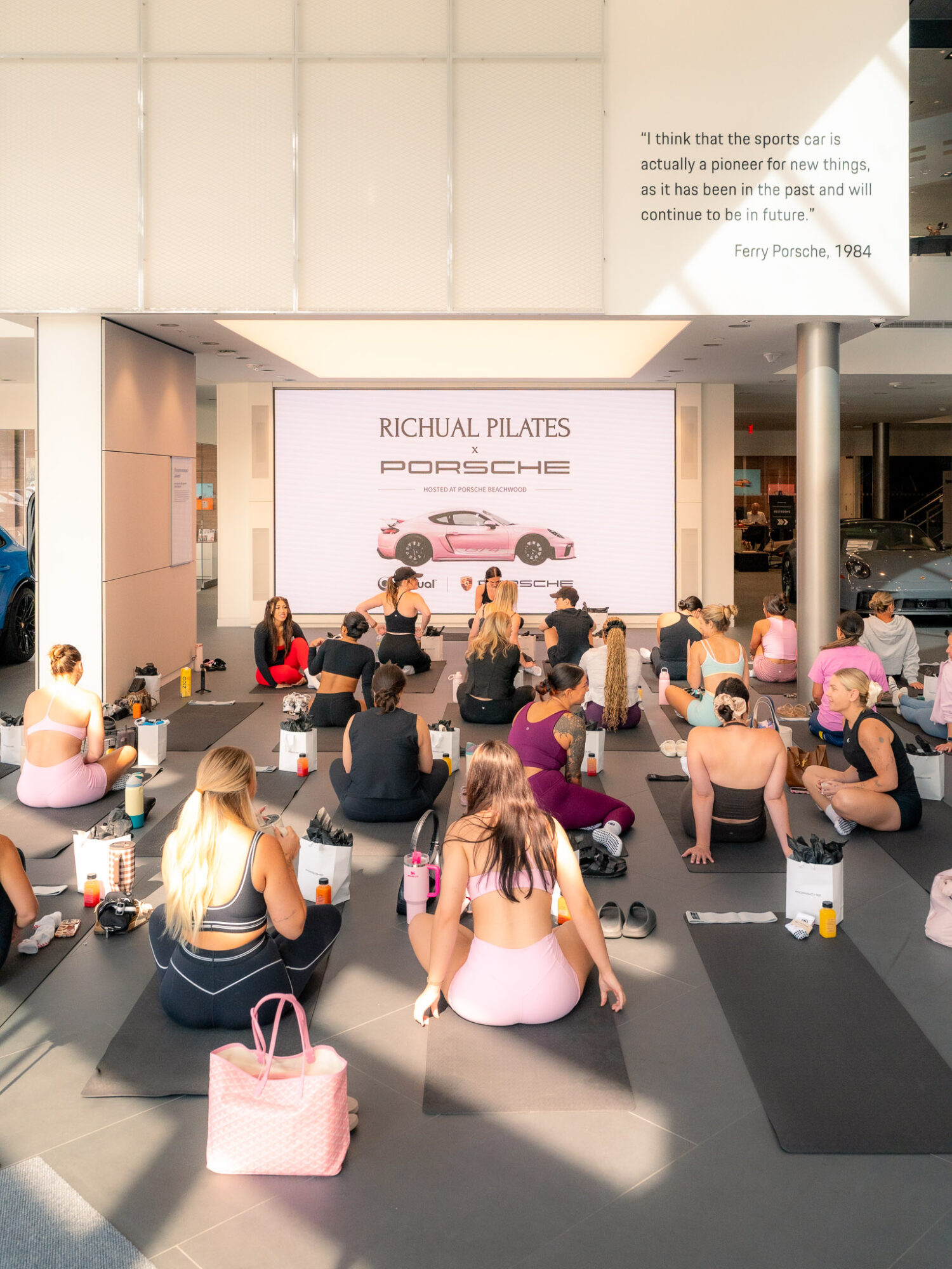 Group of people sitting on the floor facing a large screen displaying a pink Porsche car at an indoor event.