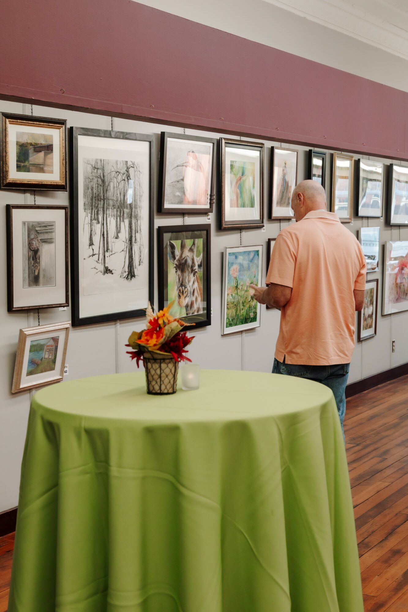 Elderly man in peach shirt viewing framed artwork on gallery wall, green table with flower arrangement in foreground.