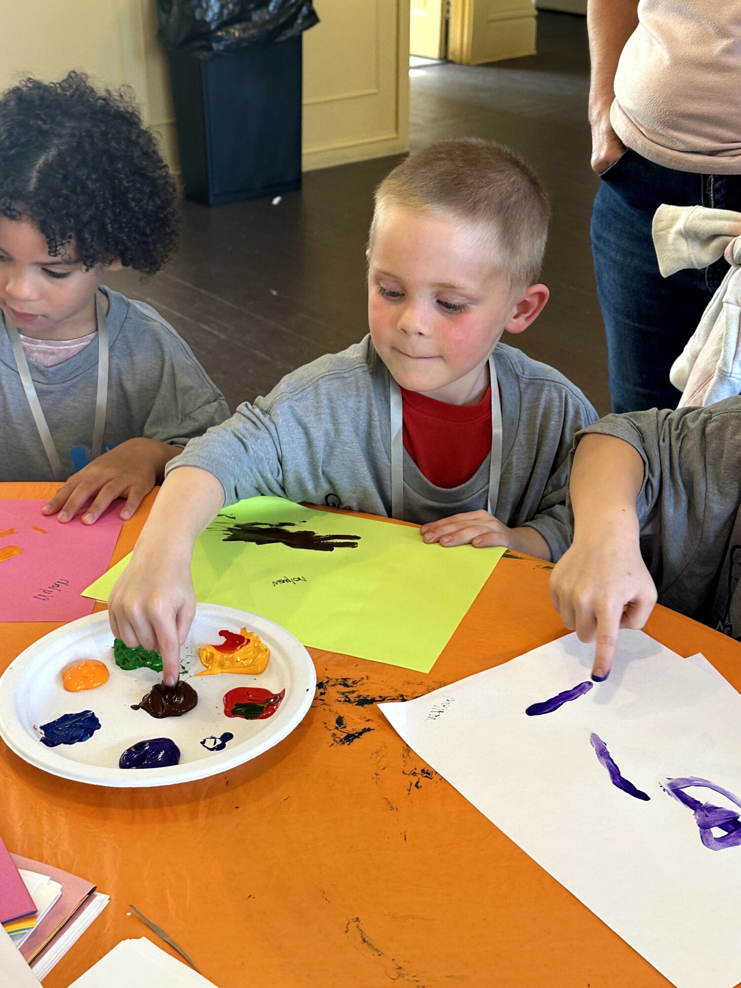 Children sit at a table painting with colorful paints, focusing on their artwork, with a person standing nearby in the background.