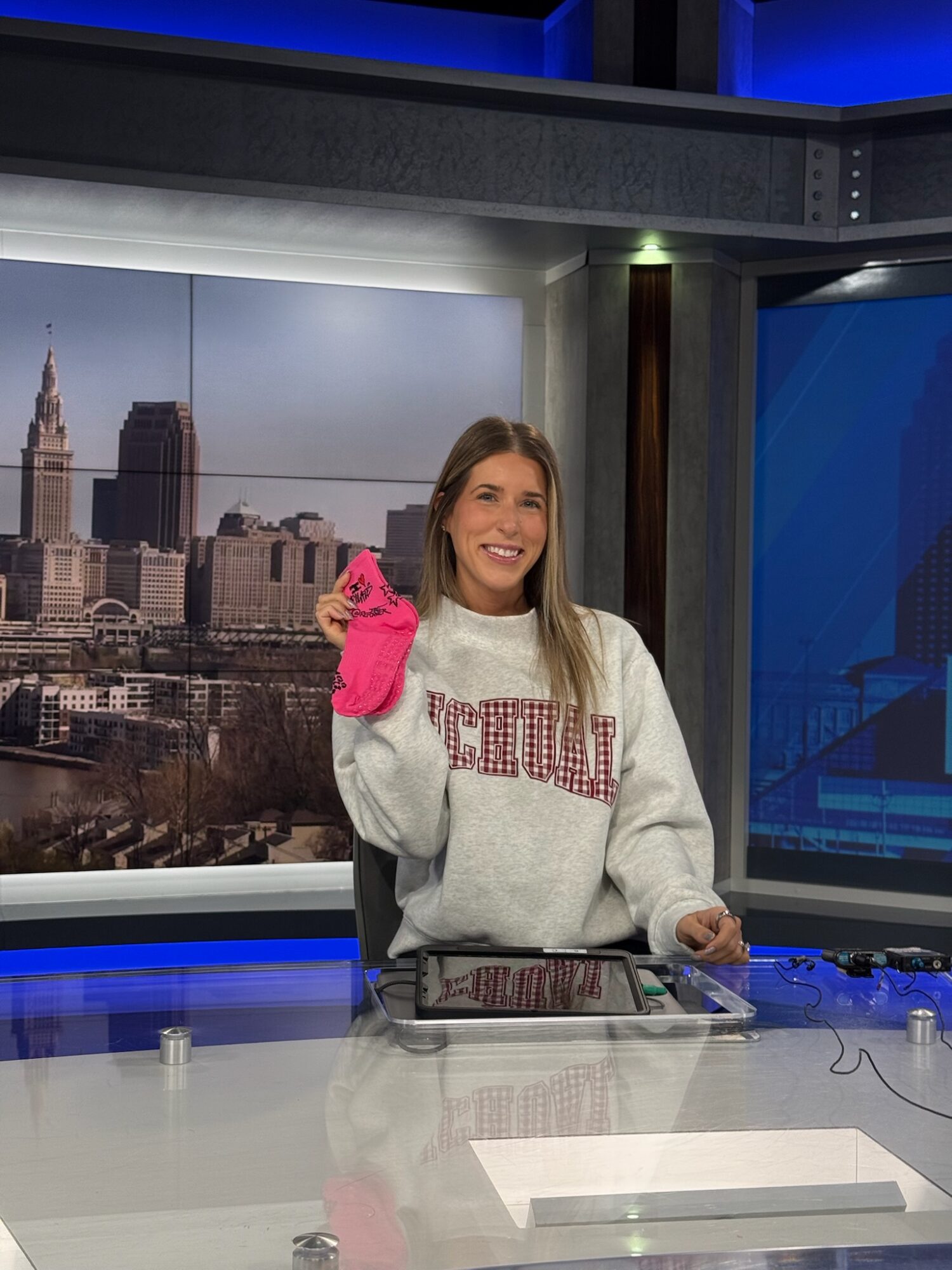 Woman in a gray sweatshirt smiling and waving in a news studio with cityscape background.