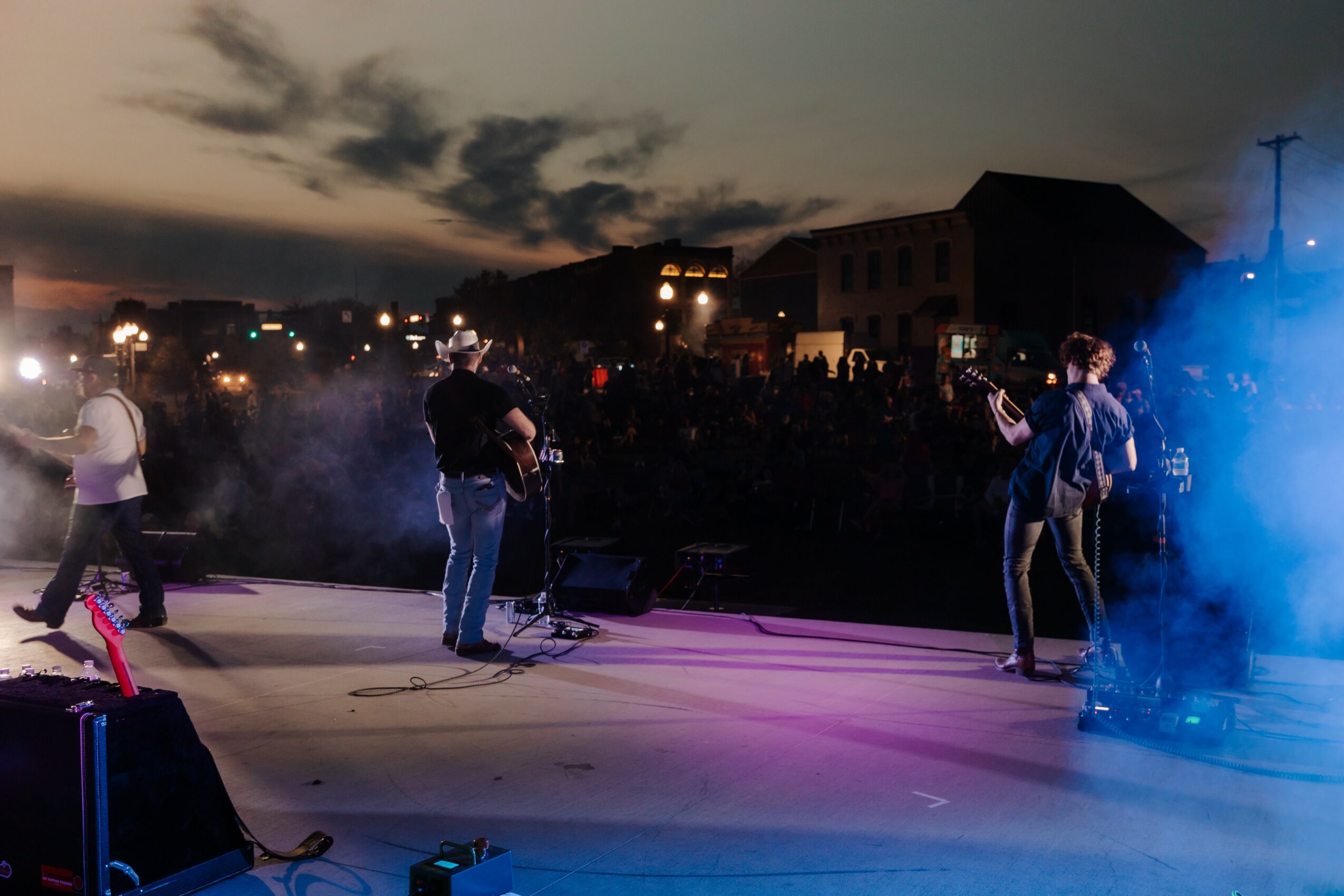 People on an outdoor stage with smoke and colorful lighting, city buildings in the background during evening.