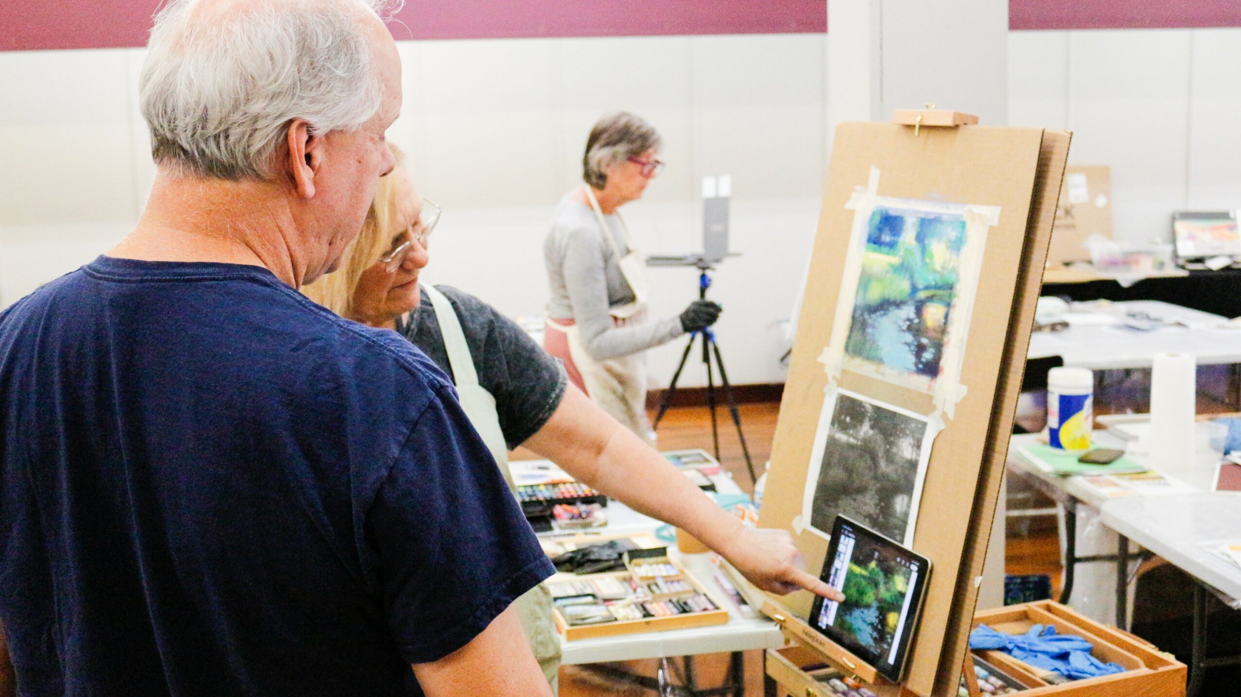 People viewing artwork at an indoor art exhibition, with paintings displayed on easels and tables in the background.