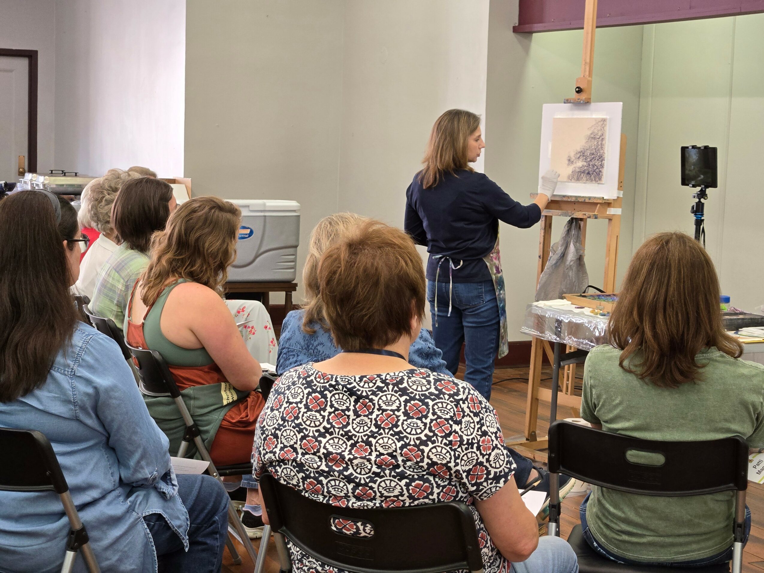 Artist demonstrating painting techniques to a group of people in an indoor setting.