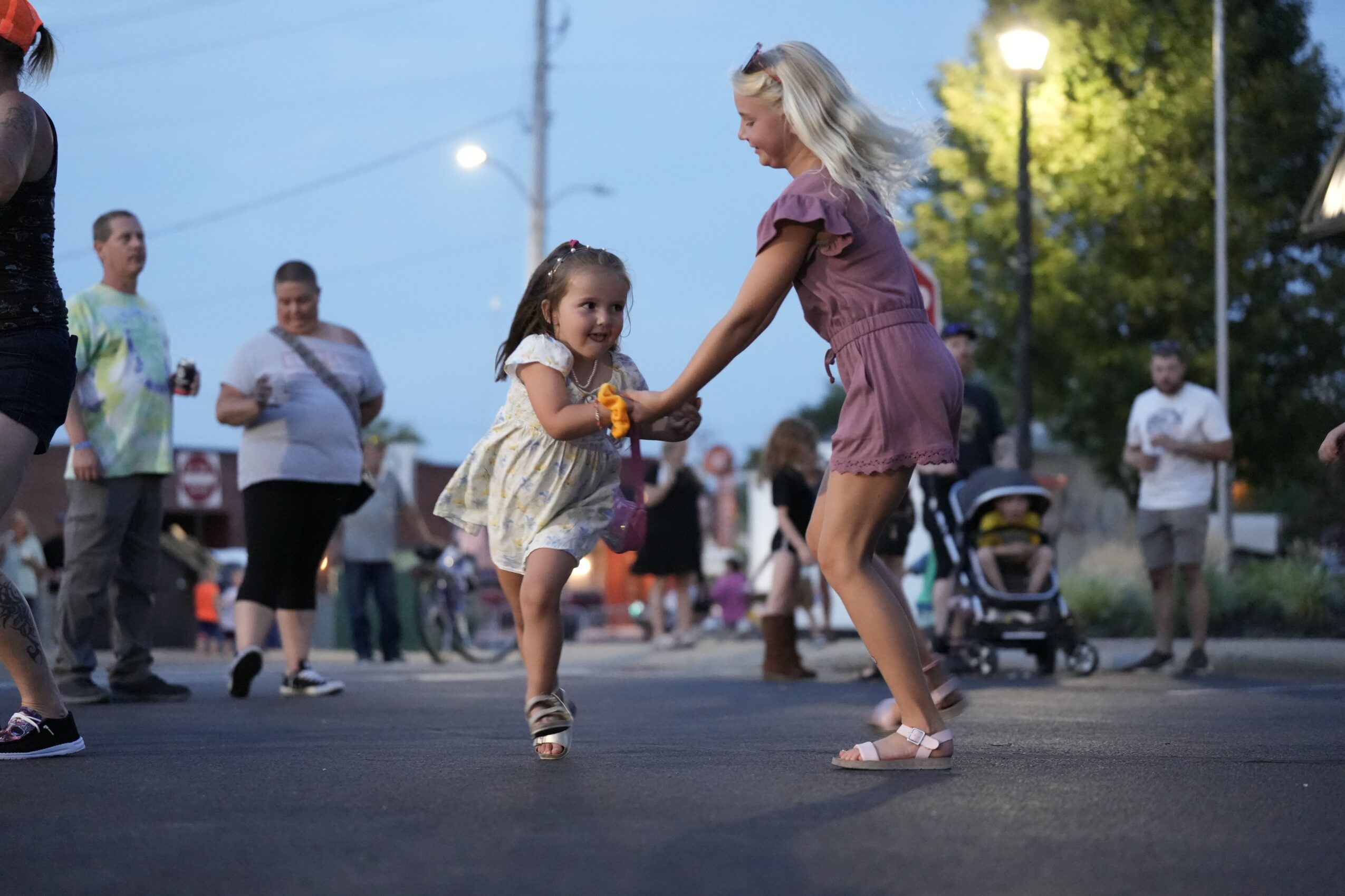 Two young girls dance and hold hands in a lively outdoor gathering during evening, with people and trees in background.