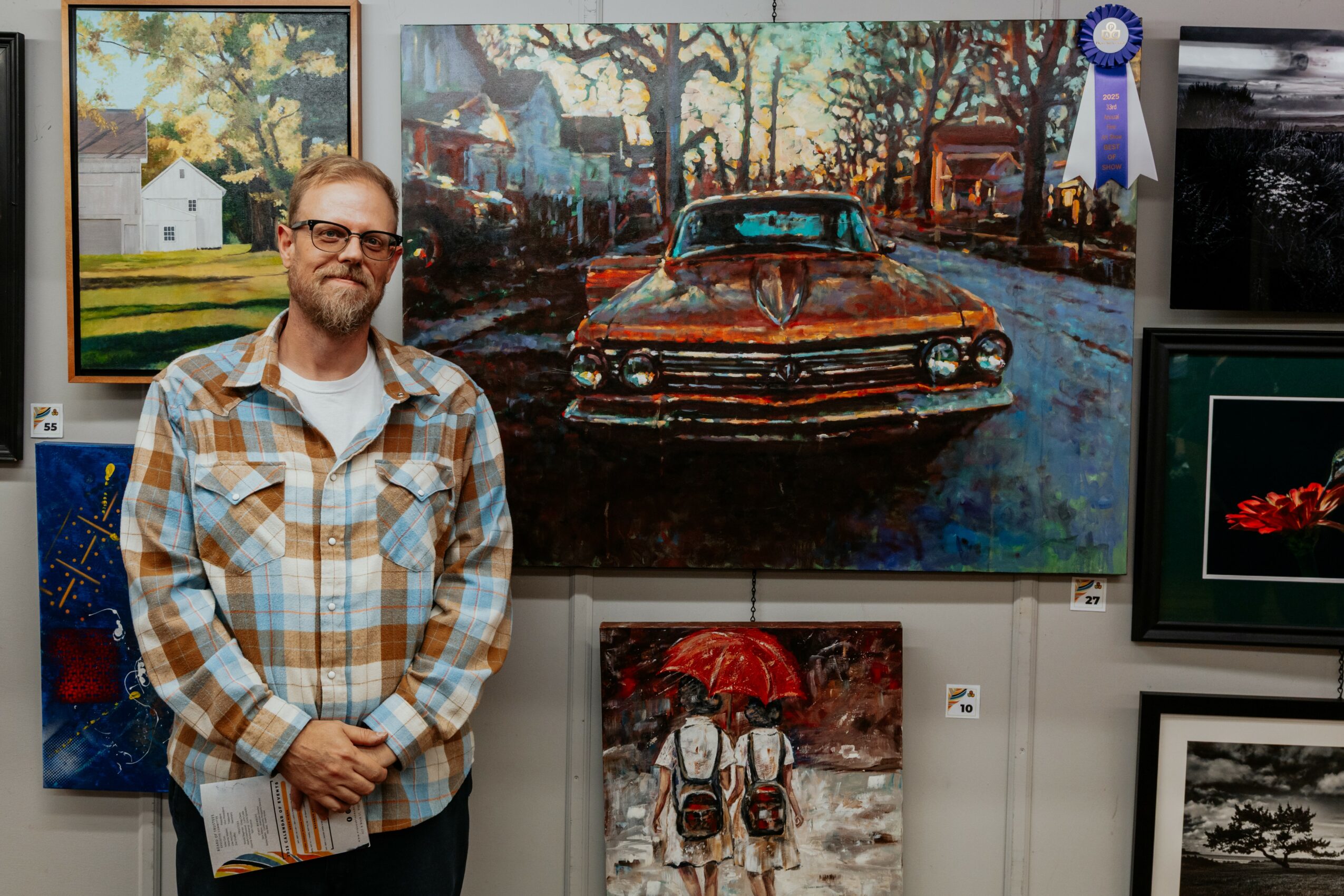 Man with glasses and plaid shirt standing in front of a large painting of a vintage car on a street.