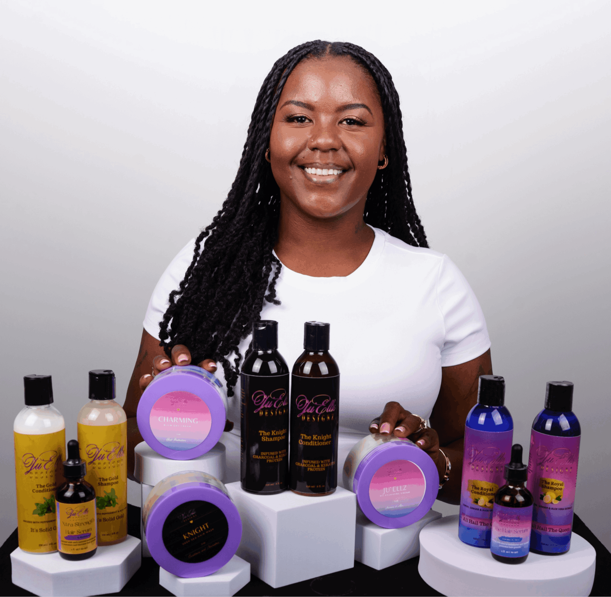 Smiling woman with long braids displays skincare products on white platforms, background is plain white.