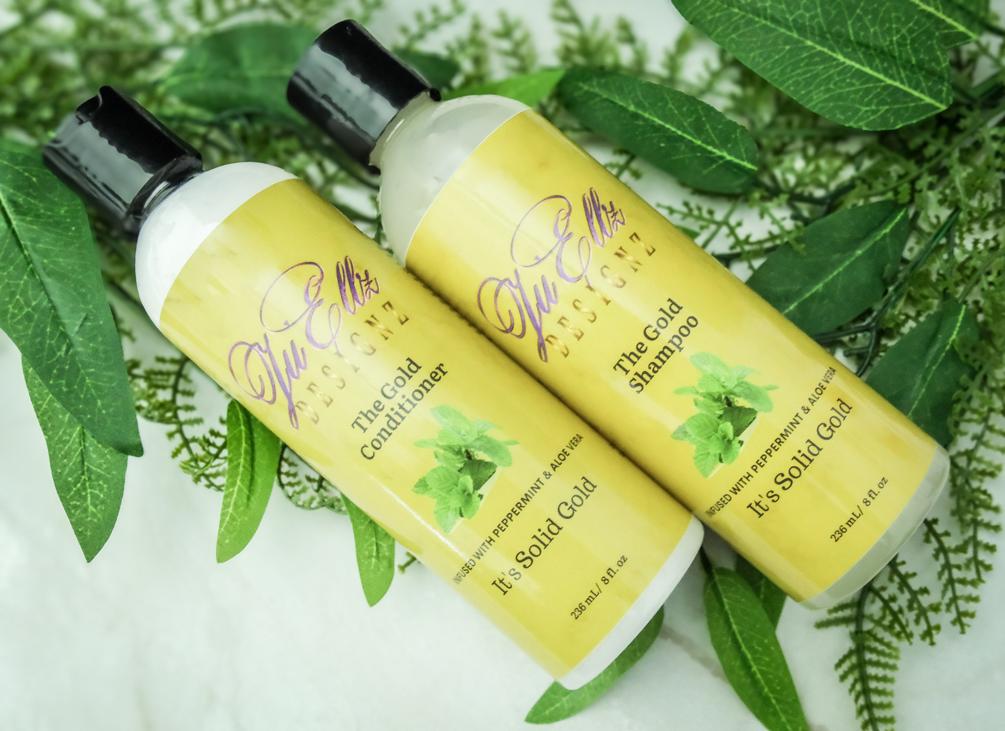 Two yellow bottles of tea tree oil shampoo with black caps, surrounded by green leaves, on a white surface.