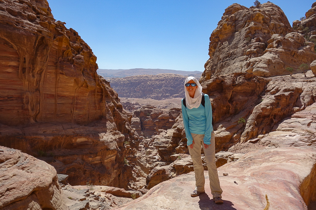 Person standing between rocky canyon walls wearing sunglasses, a light blue jacket, and beige pants, with a desert landscape in background.