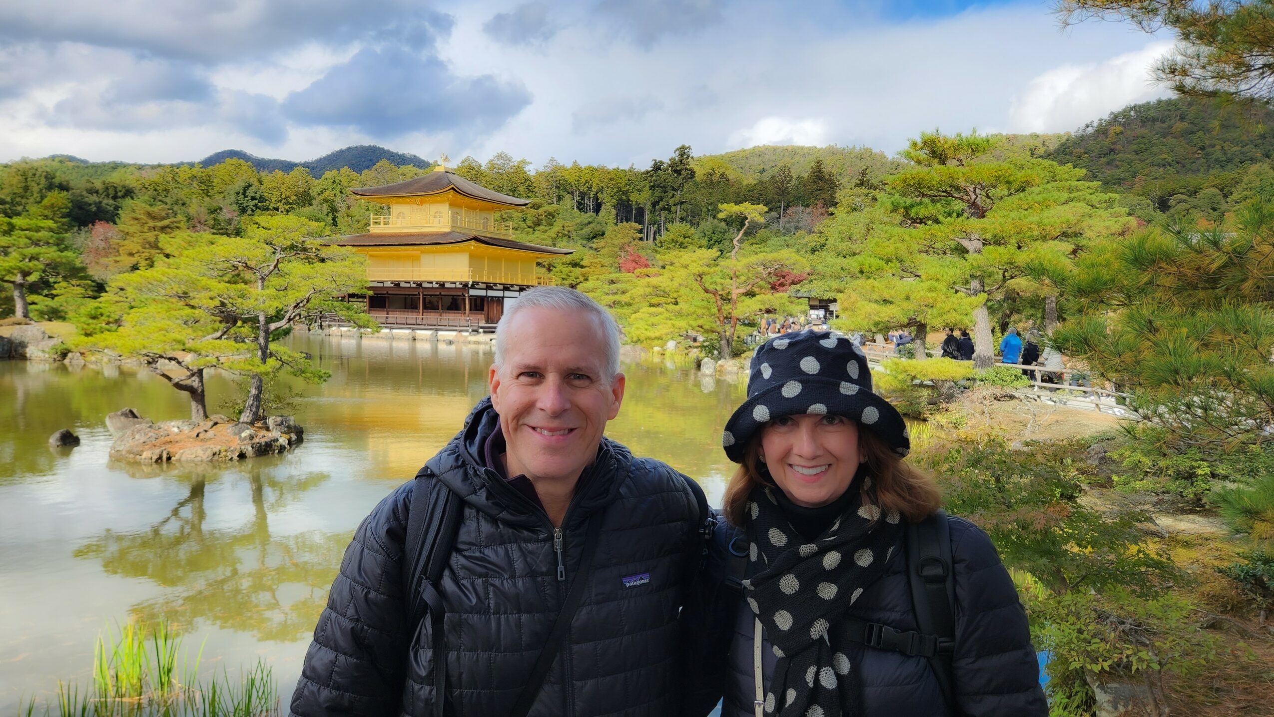 Two people smiling outdoors near a pond with trees and a traditional building in the background.