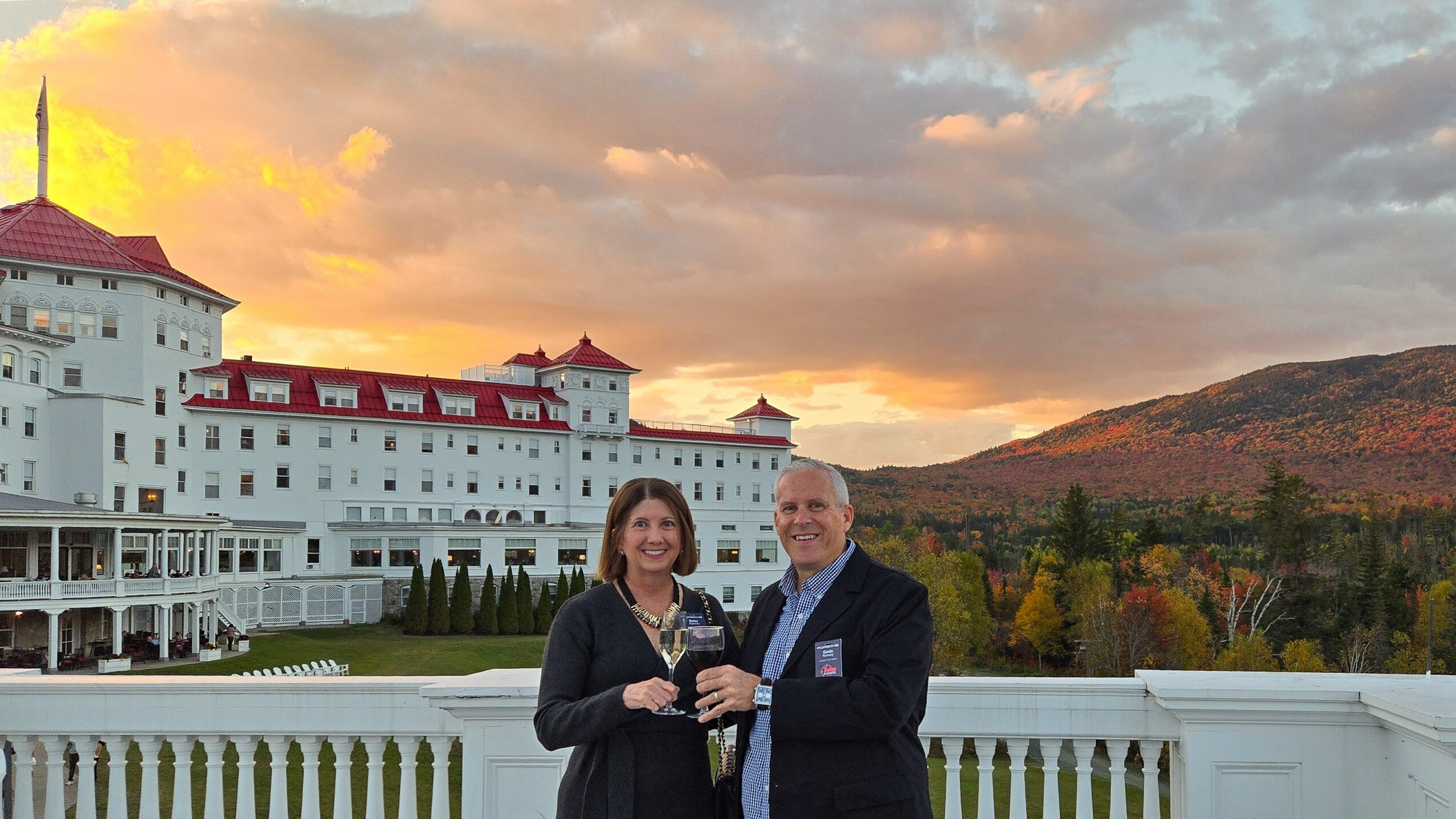 Two people standing outdoors with a large white building and hills in the background, during sunset.