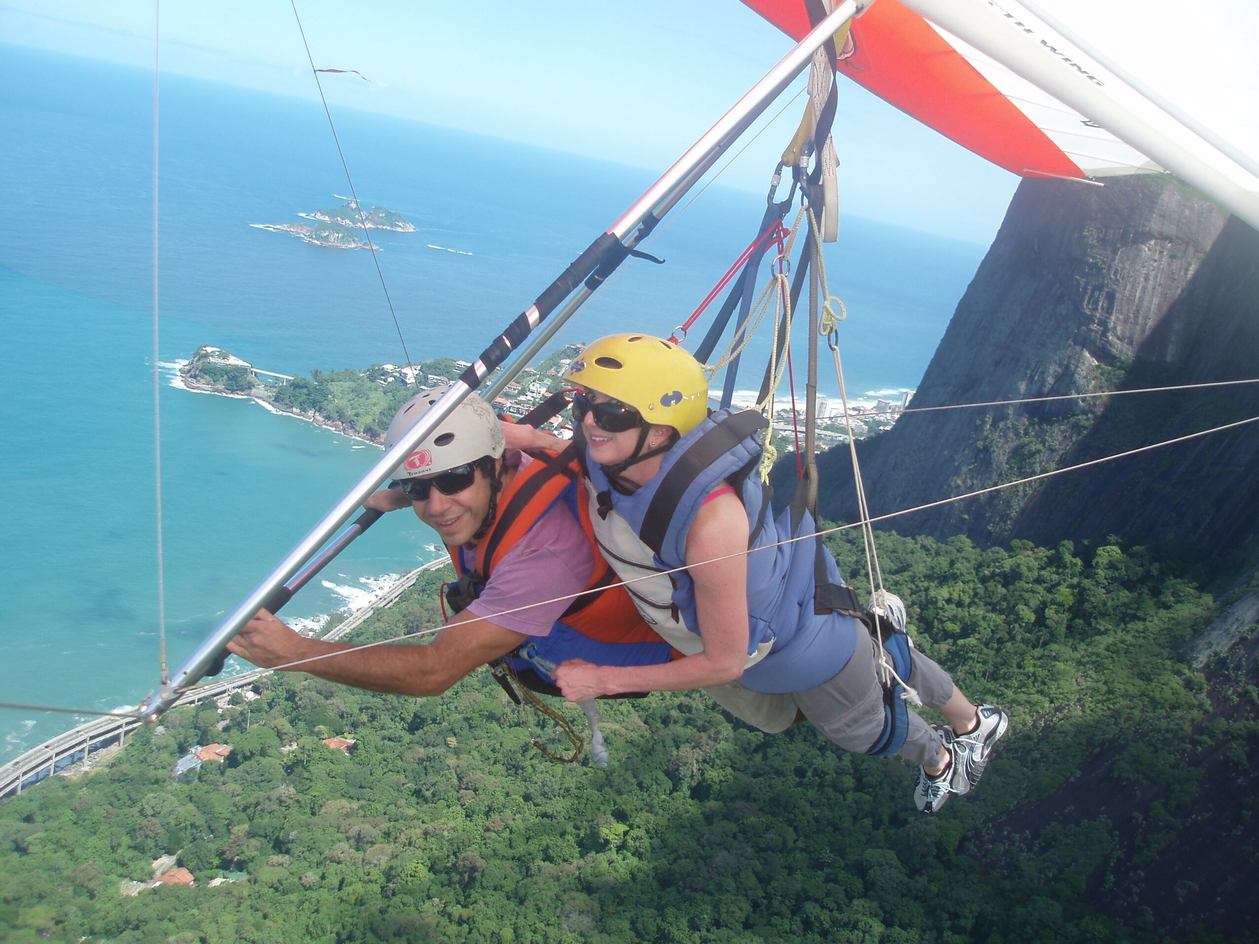 Two people hang from a hang glider high above green landscape and water, smiling and wearing helmets and harnesses.