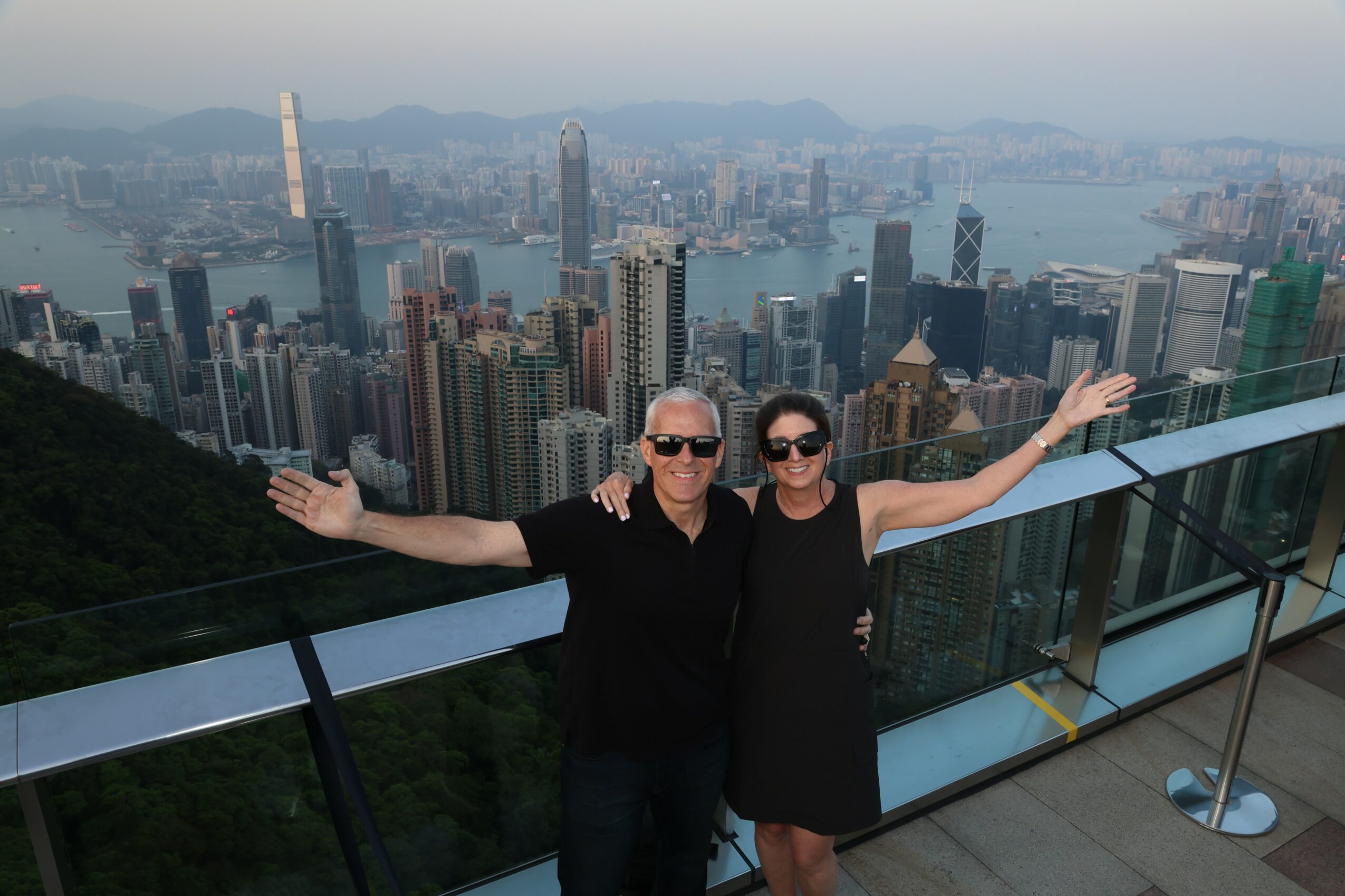 Two people stand on a balcony with a city skyline and harbor in the background, arms outstretched.