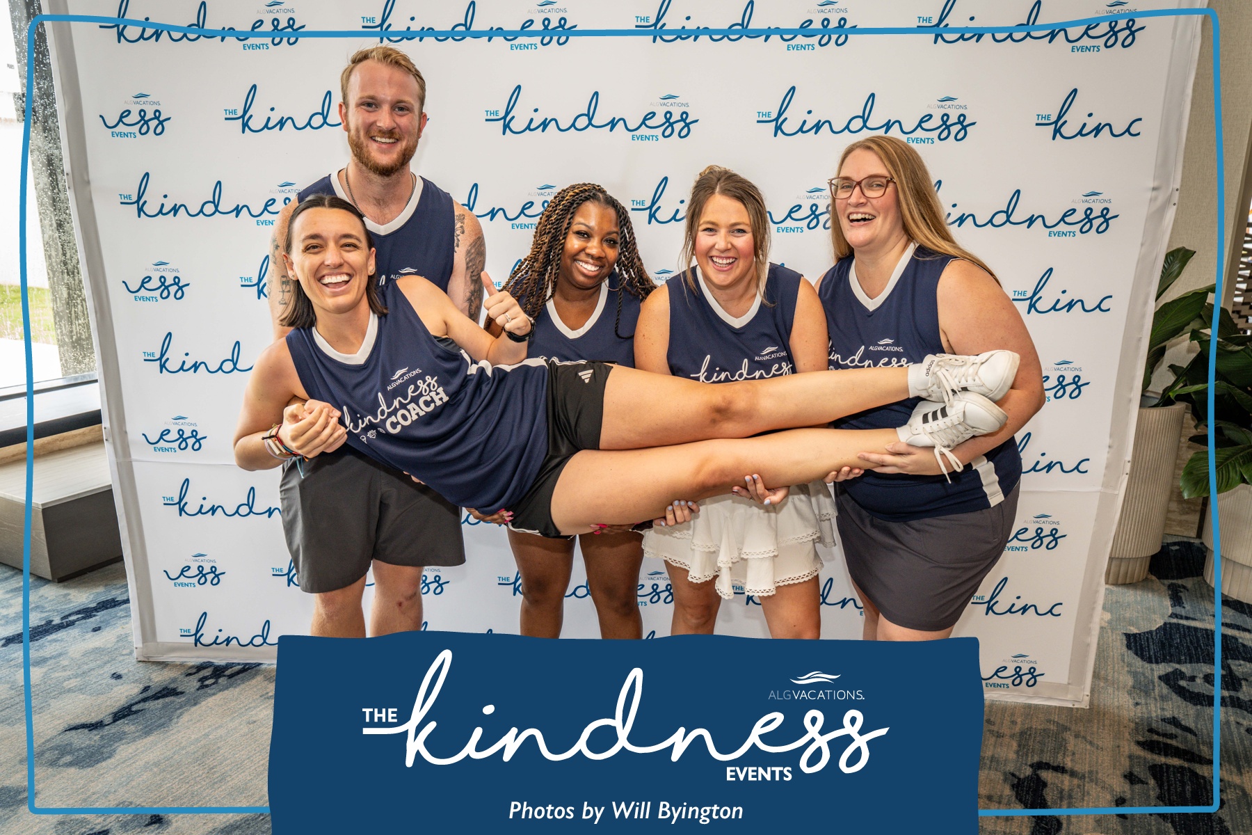 Five people in matching navy shirts pose with one person lying horizontally, smiling, in front of a backdrop with the word 'kindness' repeated. Four women and one man, all smiling, hold the person horizontally. The person being held is wearing white shoes and a white skirt. The backdrop has a blue banner reading 'The Kindness Events.'