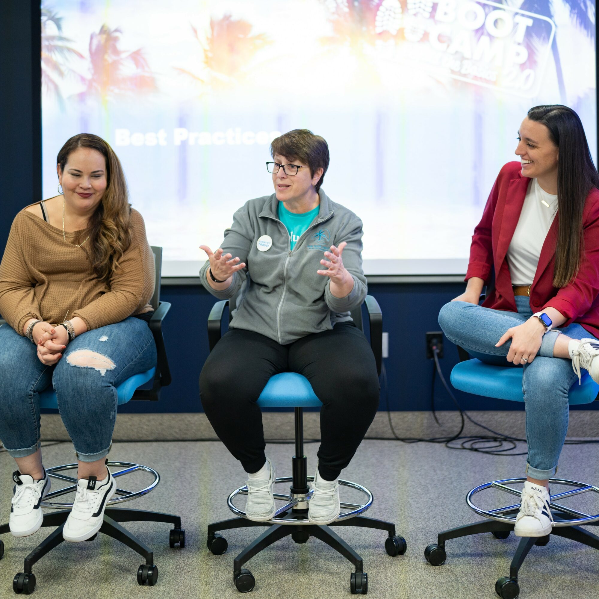 Three women sitting on chairs and engaging in conversation in a room with a screen behind them, smiling and gesturing.