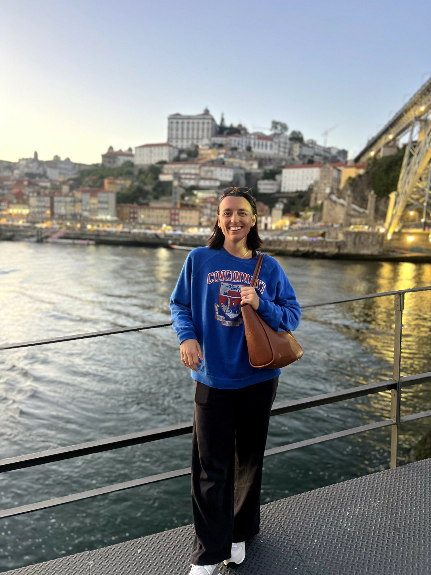 Woman standing by river with cityscape and bridge in background, smiling, holding a brown bag, wearing a blue sweatshirt and black pants.